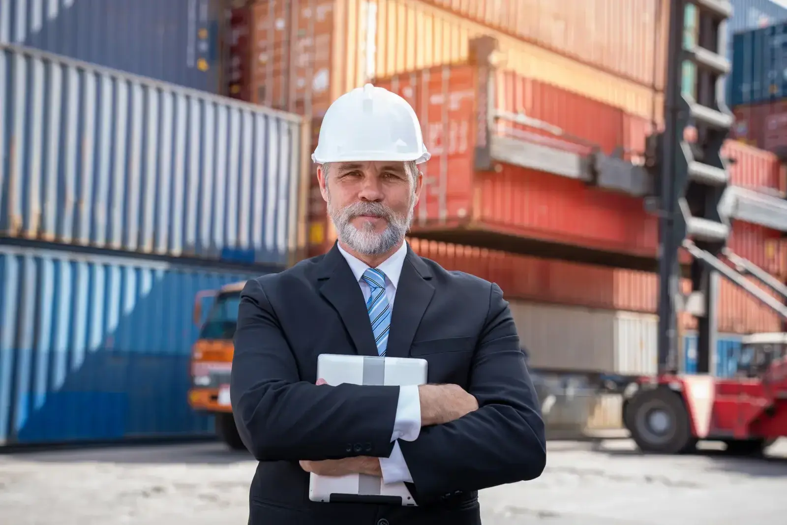 A customs clearance agent in a suit and safety helmet holding a notebook in front of several cargo containers.