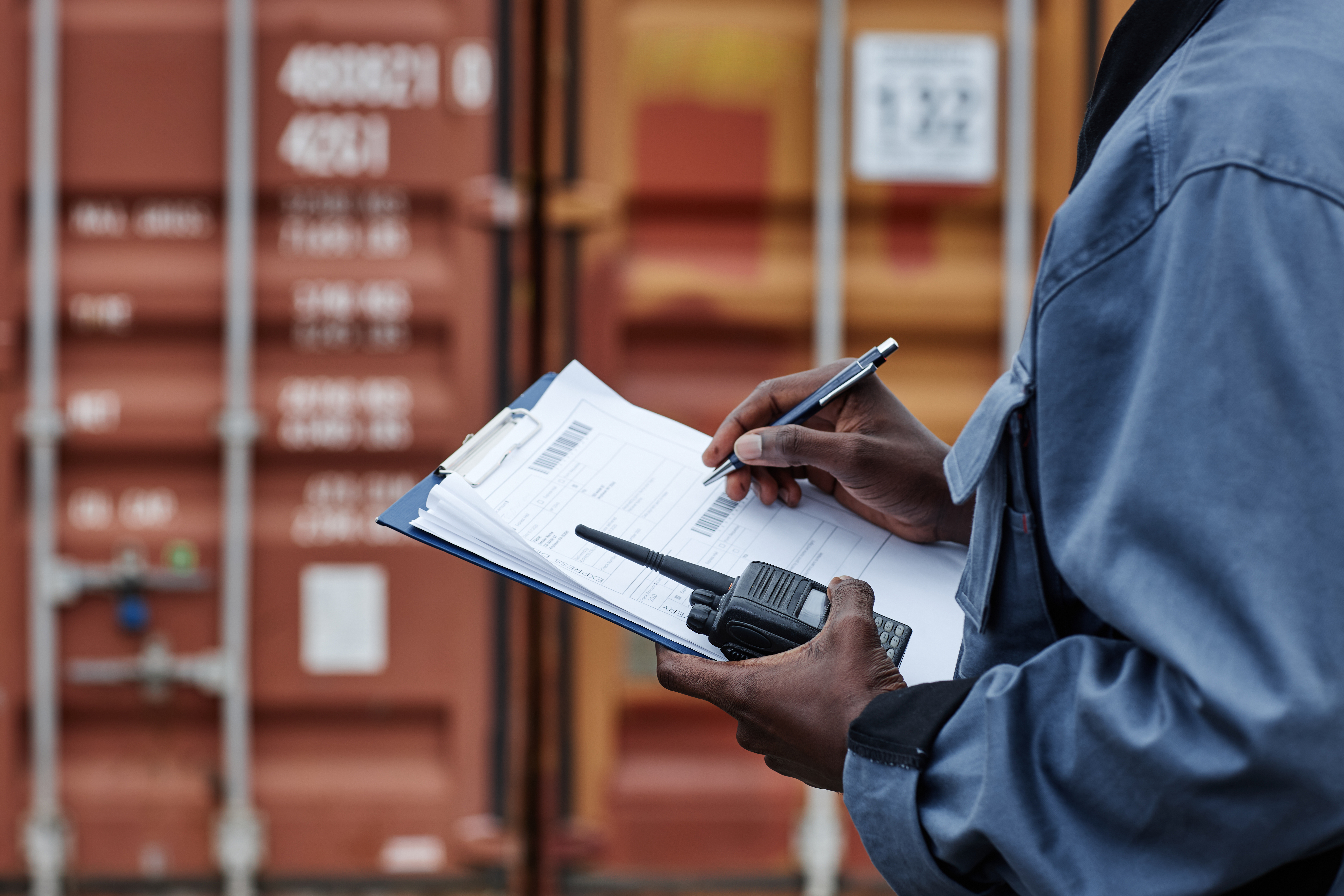 Port logistics worker reviewing customs clearance information on a clipboard beside a shipping container.